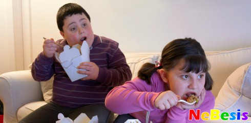 Overweight Brother and Sister Sitting Side by Side on a Sofa Eating Takeaway Food and Watching the TV